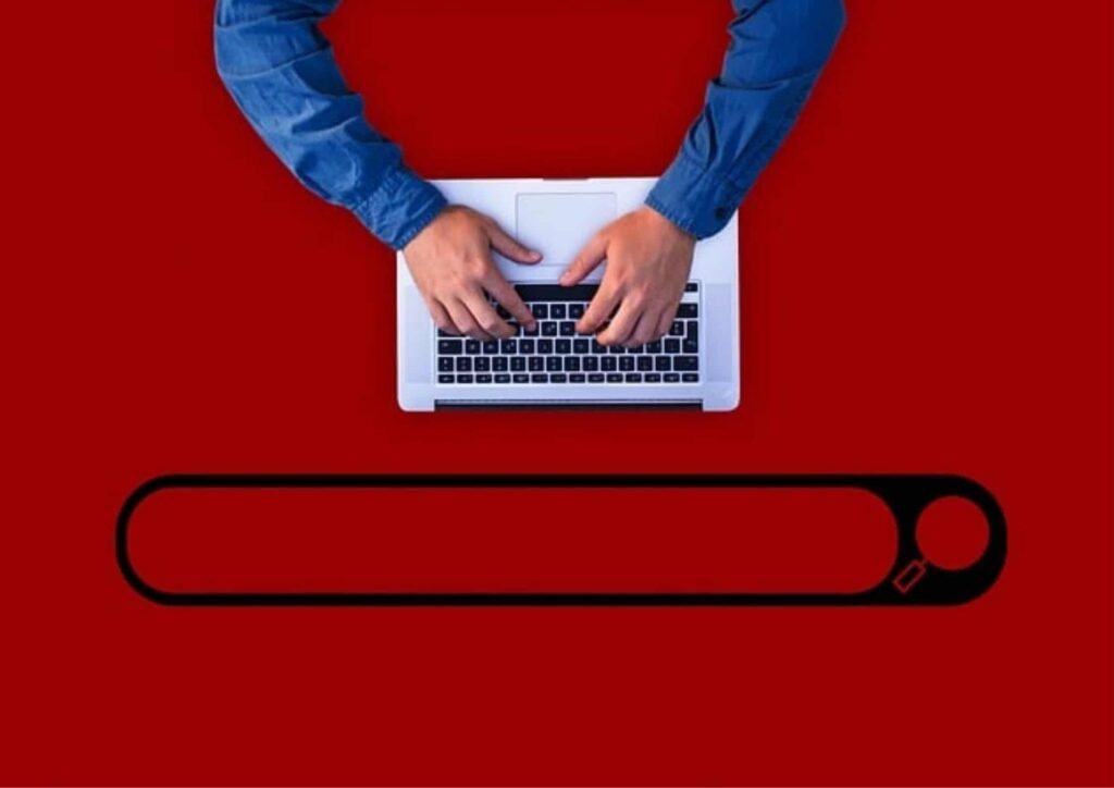 Top-down view of hands typing on a laptop above a search bar on a red background, representing AI brand visibility tools and search monitoring used to track brand presence and search performance online.